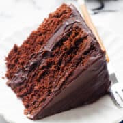 A slice of moist devil's food cake with rich chocolate frosting on a white scalloped plate, a fork resting beside it on a folded dark blue napkin, all presented on a marble background.