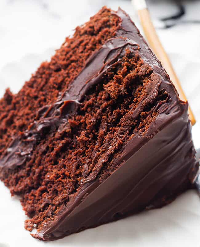 A slice of moist devil's food cake with rich chocolate frosting on a white scalloped plate, a fork resting beside it on a folded dark blue napkin, all presented on a marble background.