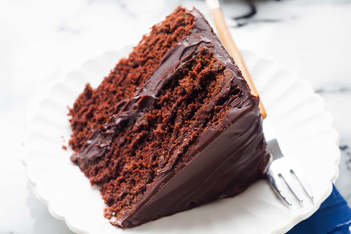 A slice of moist devil's food cake with rich chocolate frosting on a white scalloped plate, a fork resting beside it on a folded dark blue napkin, all presented on a marble background.