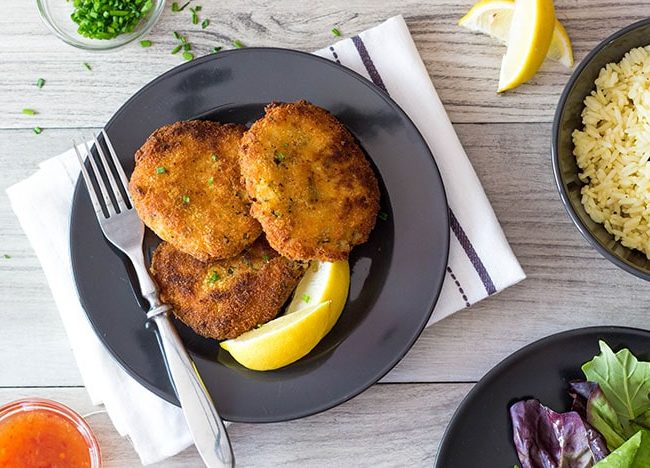 the crab cakes on a plate with lemon wedges and a dish of rice and a salad bowl in the background