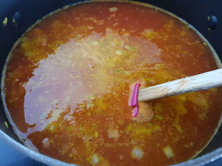 A pot of the soup with the stock and tomatoes added being mixed by a mixing spoon