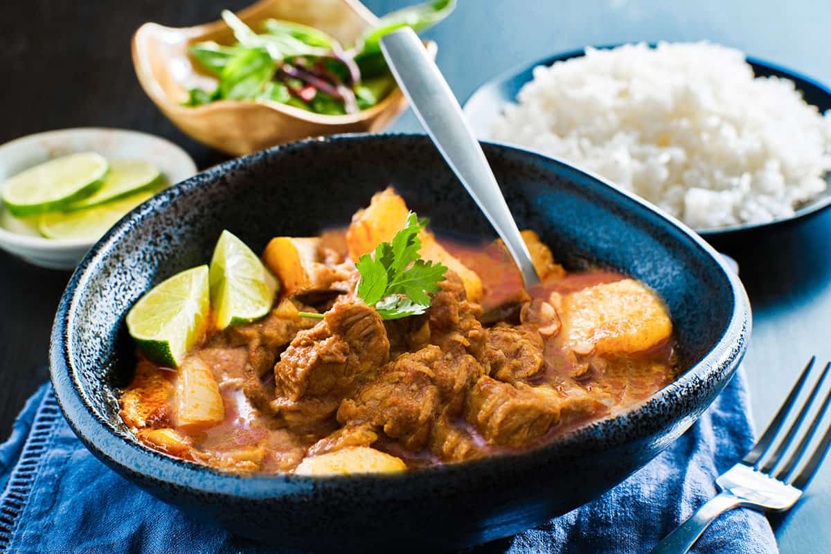 A dish of massaman curry with a bowl of white rice and limes in the background