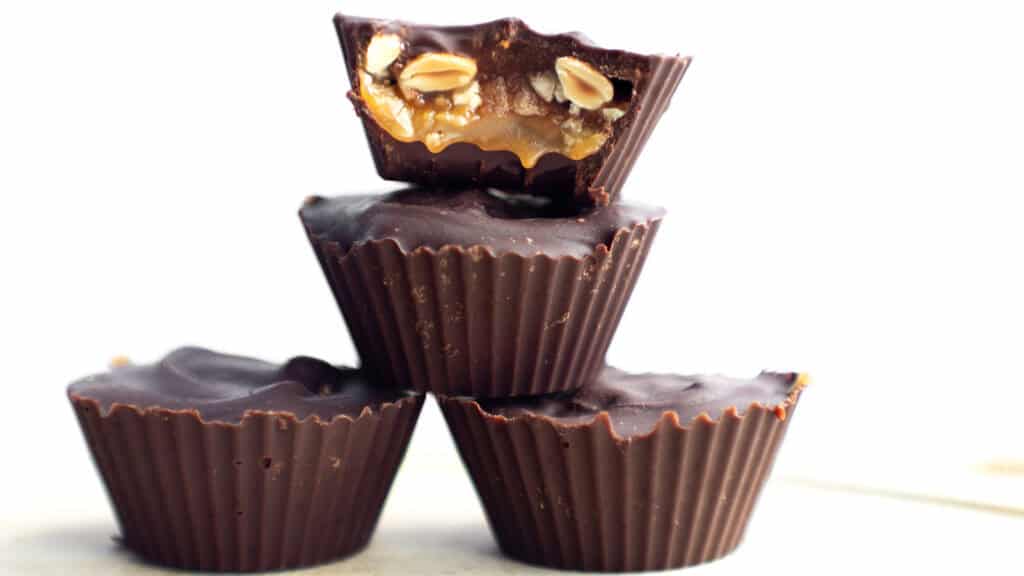 Three homemade Snicker Cups are stacked on top of each other. The top cup is bitten to reveal a filling of caramel, nuts, and possibly nougat. The cups have a dark chocolate outer layer and are displayed against a plain, white background.