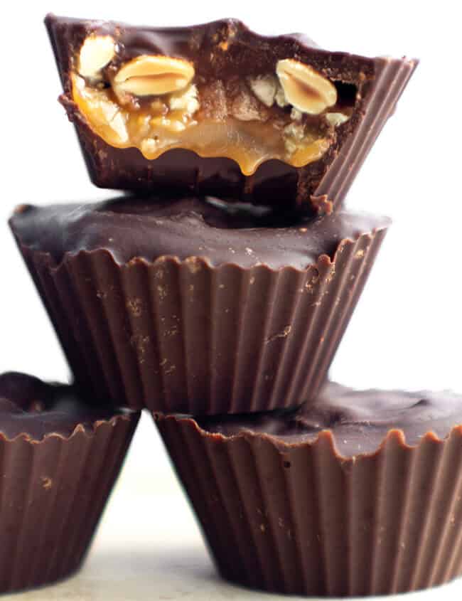 Three homemade Snicker Cups are stacked on top of each other. The top cup is bitten to reveal a filling of caramel, nuts, and possibly nougat. The cups have a dark chocolate outer layer and are displayed against a plain, white background.