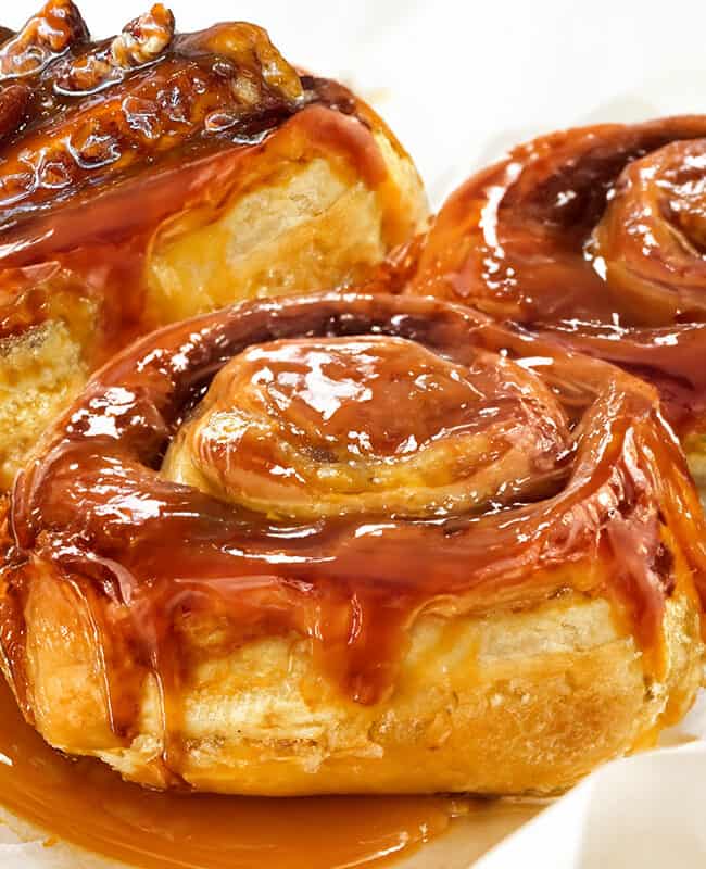 Three freshly made sticky buns covered in caramel sauce and one topped with chopped pecans.