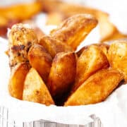 A serving of golden-brown potato wedges in a metal basket lined with white parchment paper. The wedges are crispy, and a tray of more potato wedges is blurred in the background.