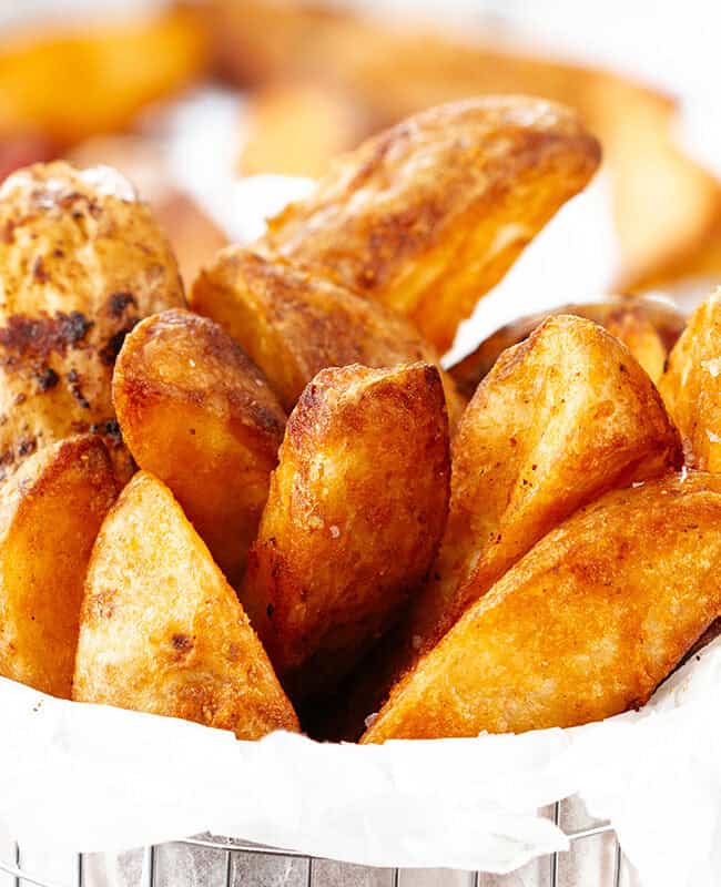 A serving of golden-brown potato wedges in a metal basket lined with white parchment paper. The wedges are crispy, and a tray of more potato wedges is blurred in the background.