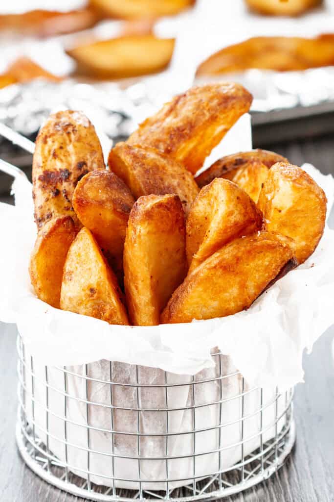 A metal basket lined with white parchment paper is filled with golden-brown potato wedges. These crispy, seasoned potato wedges stand upright and are piled high in the basket. In the background, additional food items are slightly out of focus on a foil-lined tray.