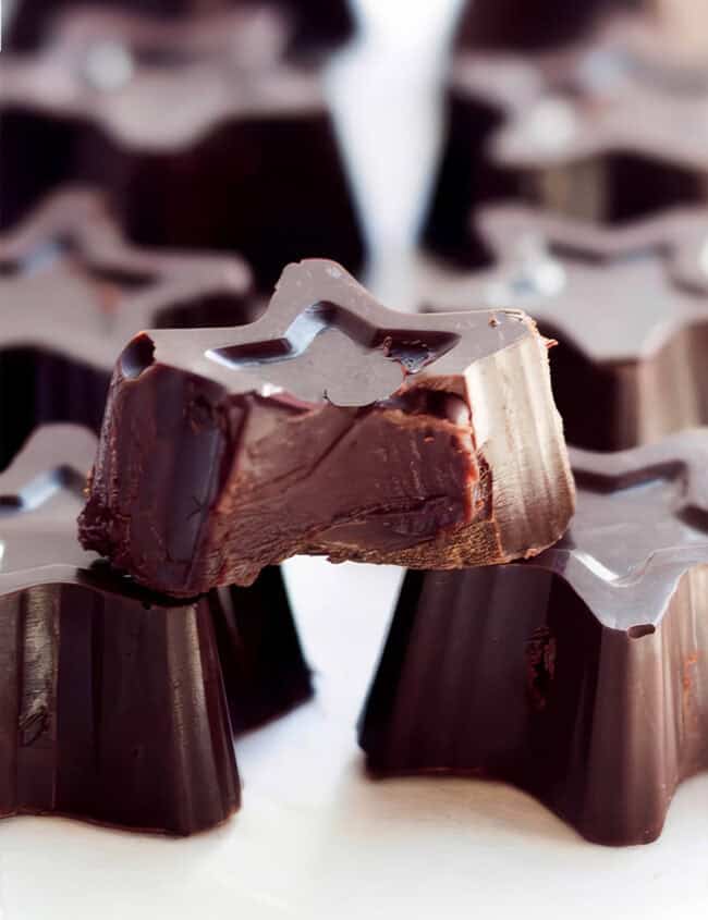 Close-up of star-shaped dark chocolate truffles, one with a bite taken out of it revealing a creamy filling inside. The chocolates are arranged in a cluster on a light surface, with a blurred background highlighting the focus on the bitten piece.