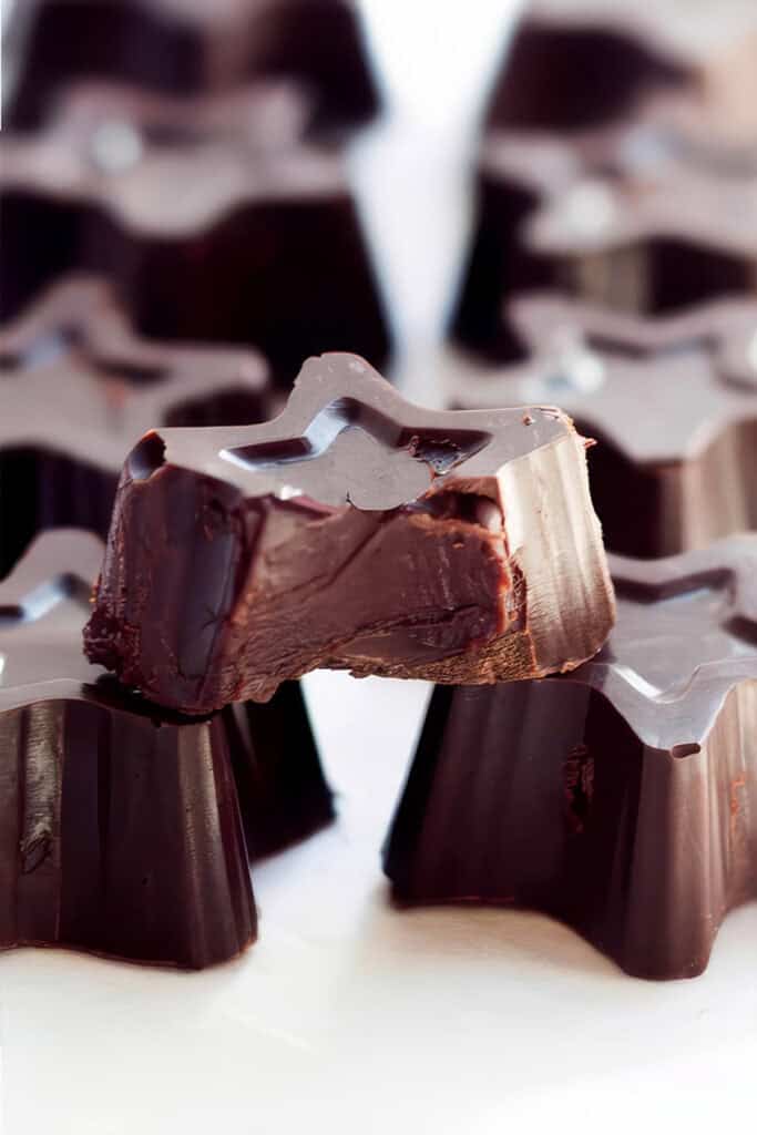 Close-up of star-shaped dark chocolate truffles, one with a bite taken out of it revealing a creamy filling inside. The chocolates are arranged in a cluster on a light surface, with a blurred background highlighting the focus on the bitten piece.