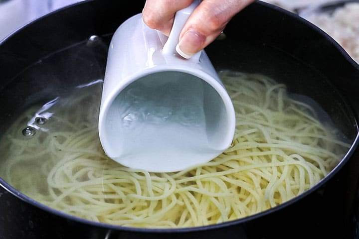 A pot of spaghetti cooking and a mug collecting some water from the pot