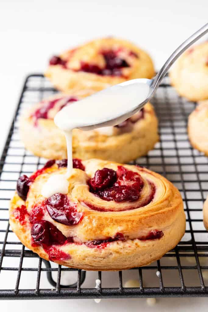 A spoon drizzles white icing onto a cherry-filled pastry roll on a cooling rack. The pastry is a burst of color, combining vibrant red cherries with hints reminiscent of iced cranberry and orange swirl scones. More rolls rest invitingly in the background against a plain white surface.