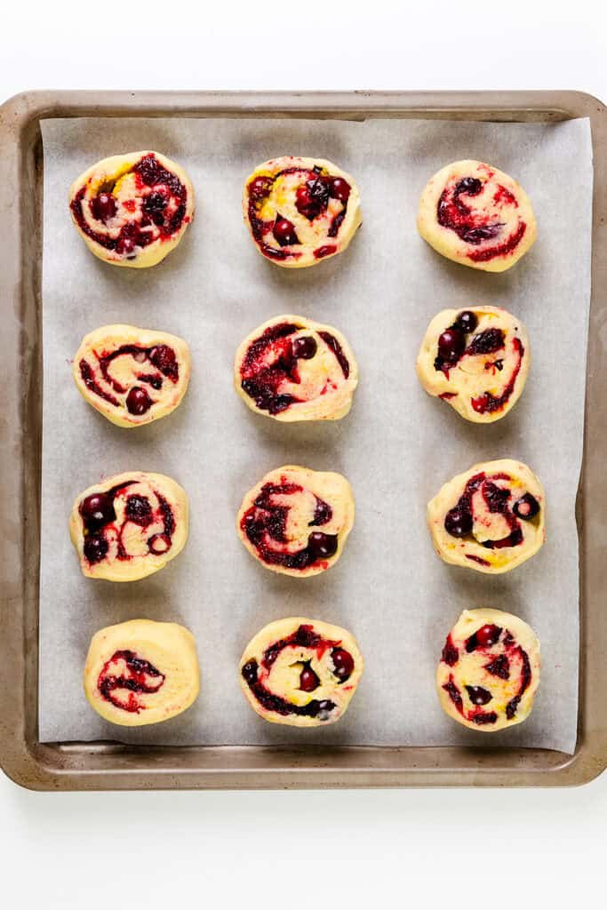 Twelve unbaked pinwheel pastries with a berry filling are arranged on a parchment-lined baking sheet. The vibrant red and dark fruit swirls evoke the delightful pattern of iced cranberry and orange swirl scones against the light background of the tray.