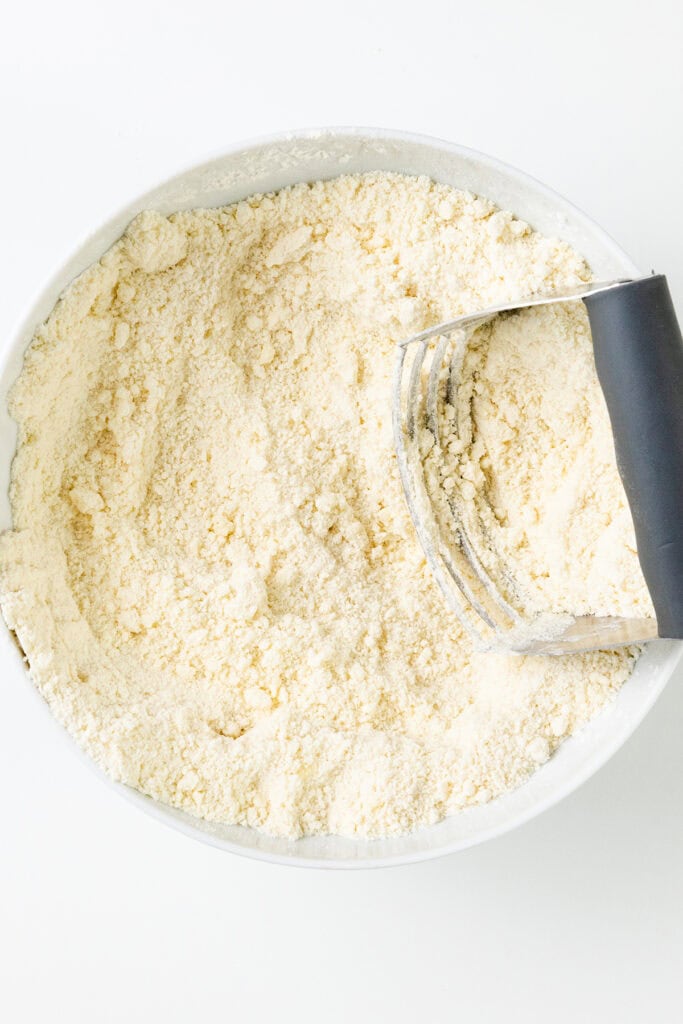 A white bowl filled with a crumbly flour mixture, perfect for crafting delectable Iced Cranberry & Orange Swirl Scones, features a pastry cutter with a black handle partially submerged in the mixture, resting on a light surface.