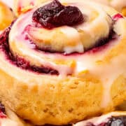 A close-up of freshly baked iced cranberry and orange swirl scones, topped with white icing and a dollop of red preserves. The scones boast a golden-brown crust, swirled with berry filling, showcasing a deliciously inviting appearance.