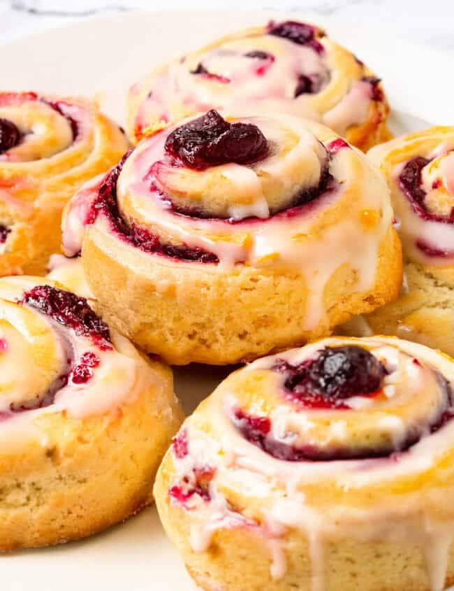 Close-up of glazed Cranberry and Orange Scones arranged on a white plate. The pastries, reminiscent of iced cranberry & orange swirl scones, are filled with cherry and topped with glossy icing, showcasing a golden-brown, flaky crust. The plate rests on a white surface.