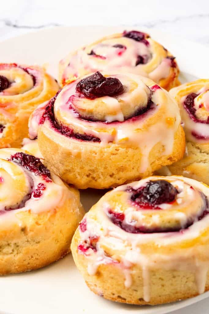 Close-up of glazed cherry rolls arranged on a white plate. The pastries, reminiscent of iced cranberry & orange swirl scones, are filled with cherry and topped with glossy icing, showcasing a golden-brown, flaky crust. The plate rests on a white surface.