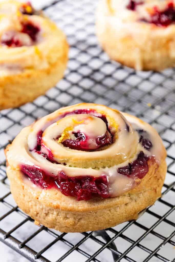 A close-up of a freshly baked cinnamon roll with swirls of glaze and a filling that hints at berries and the zest of iced cranberry & orange swirl scones, resting on a black wire cooling rack. The pastry boasts a golden-brown crust with a glossy glaze finish.