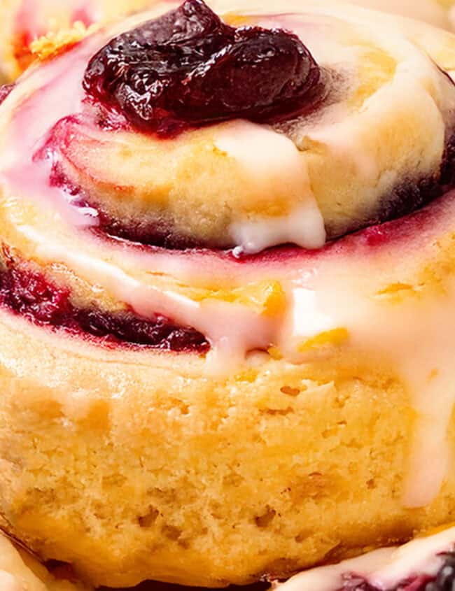 A close-up of freshly baked iced cranberry and orange swirl scones, topped with white icing and a dollop of red preserves. The scones boast a golden-brown crust, swirled with berry filling, showcasing a deliciously inviting appearance.