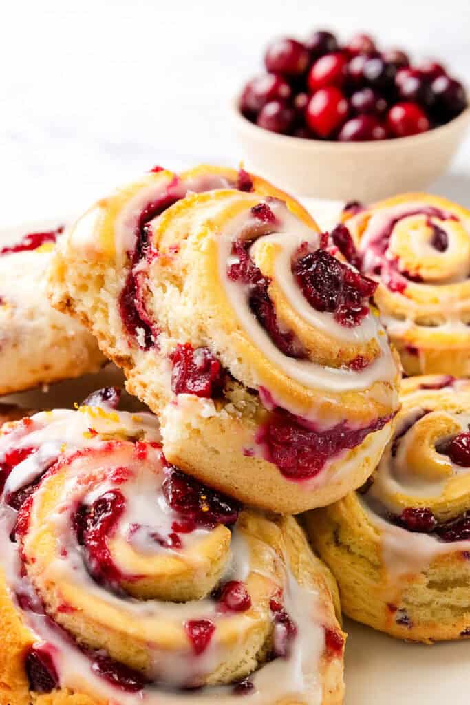 Close-up of freshly baked cranberry rolls topped with glaze, piled together on a plate. A bite is taken out of one roll, revealing the moist texture and cranberry filling reminiscent of Iced Cranberry & Orange Swirl Scones. A small bowl of cranberries is blurred in the background.