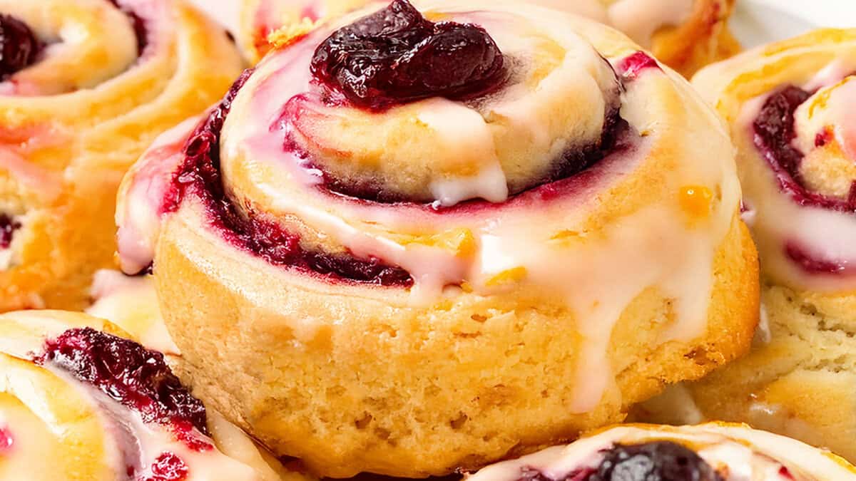 A close-up of freshly baked iced cranberry and orange swirl scones, topped with white icing and a dollop of red preserves. The scones boast a golden-brown crust, swirled with berry filling, showcasing a deliciously inviting appearance.