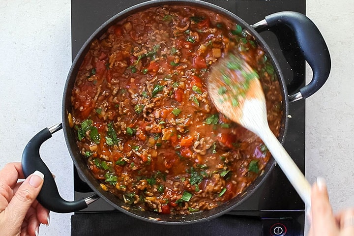 A pan with tomatoes and parsley being added to the meat, garlic and onions