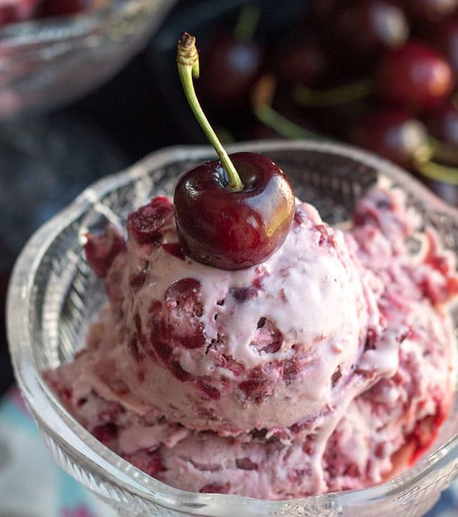 A close up view of the cherry ice cream in a glass bowl topped with a cherry and more cherries in the background