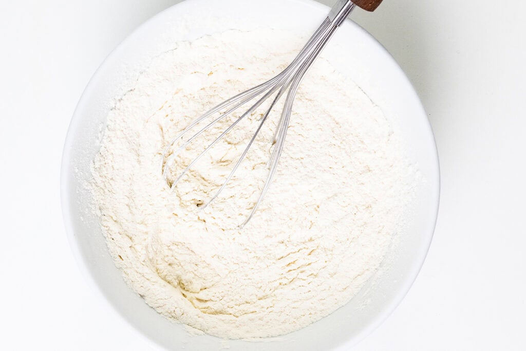 A white bowl filled with flour and a whisk with a wooden handle on top, ready to craft a delicious chocolate chip caramel cake. The finely textured flour is spread evenly, set against a plain, light background.