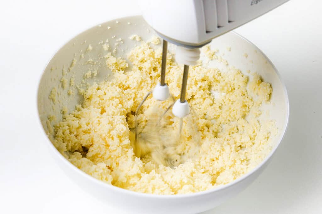 An electric mixer is blending a creamy mixture in a white bowl, likely preparing a dough or batter for a Chocolate Chip Caramel Cake. The mixture has a light, fluffy texture and pale yellow color, suggesting it's in the early stages of combining ingredients like butter and sugar.