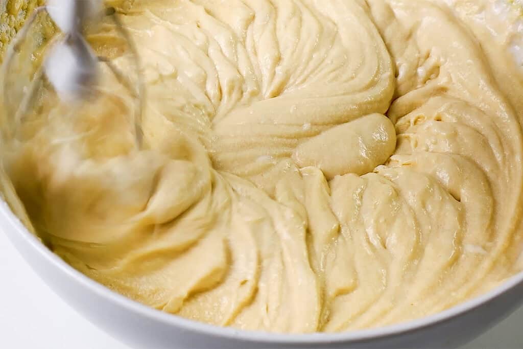 A close-up of creamy, beige cake batter with hints of chocolate chip caramel swirling in a white bowl. An electric mixer is blending the batter, creating mesmerizing patterns on the surface.