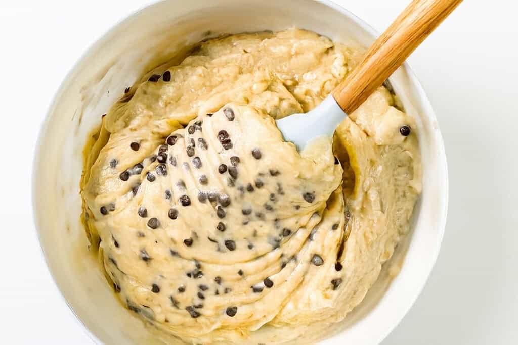 A white bowl brimming with creamy cookie dough is mixed with chocolate chips, reminiscent of a delightful chocolate chip caramel cake. A wooden-handled spatula is partially submerged, showcasing the dough's thick and smooth texture.