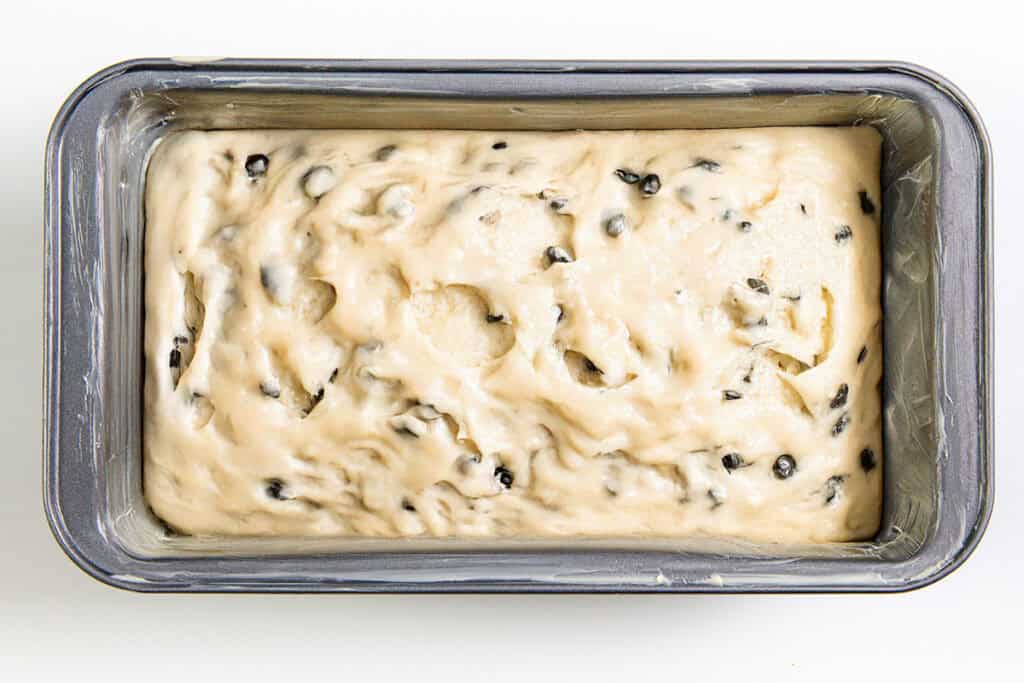 A rectangular loaf pan filled with raw bread dough, speckled with dark chocolate chips and hints of caramel, ready to be baked into a deliciously unique cake. The dough boasts a smooth, creamy texture with visible air pockets. The pan rests invitingly on a white surface.