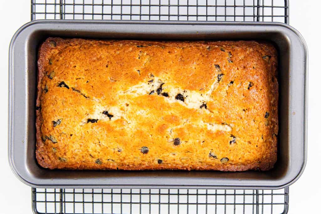 A freshly baked loaf of golden-brown banana bread with chocolate chips, reminiscent of a chocolate chip caramel cake, sits in a rectangular metal pan on a black wire cooling rack. The bread's cracked surface reveals its moist, textured interior.