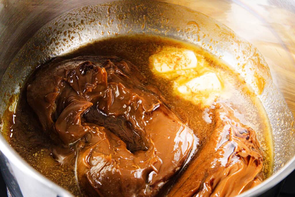 A pot containing simmering caramelized brown sauce with creamy butter melting on the side, ideal for drizzling over a Chocolate Chip Caramel Cake. The luscious, glossy texture suggests it might be dulce de leche or caramel cooking in progress.