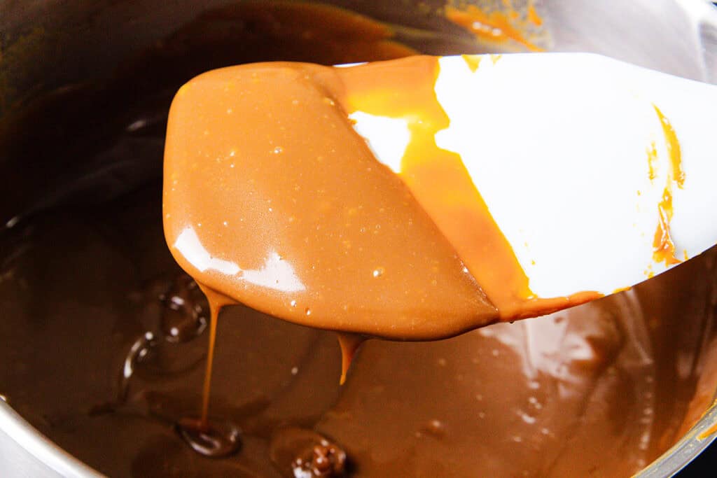 Close-up of a spatula lifting smooth, melted caramel from a pan, destined for a decadent Chocolate Chip Caramel Cake. The glossy, rich golden-brown hue adorned with bubbles hints at the perfect blend of sweetness and texture as it’s stirred to velvety perfection.