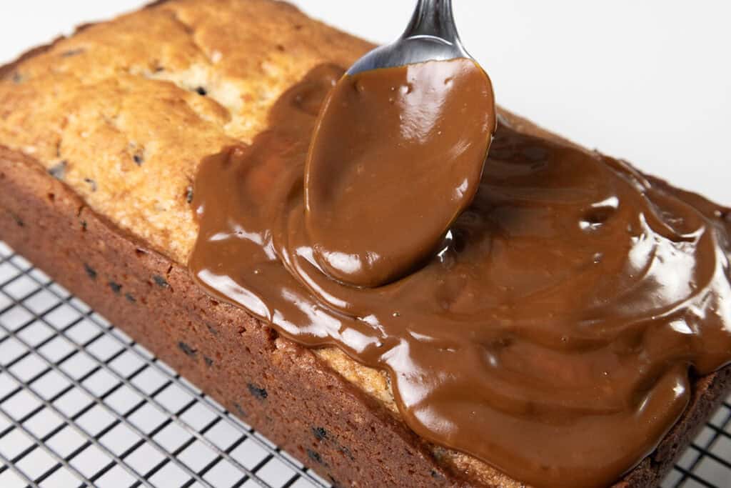 A spoon spreads glossy caramel sauce over a freshly baked chocolate chip caramel cake on a cooling rack. The cake boasts a golden-brown crust, studded with chocolate chips, and is luxuriously topped with thick, rich caramel.