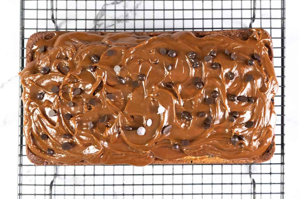 A rectangular chocolate chip caramel cake with thick, swirled chocolate frosting, topped with scattered chocolate chips. The cake rests on a wire cooling rack over a marble surface.