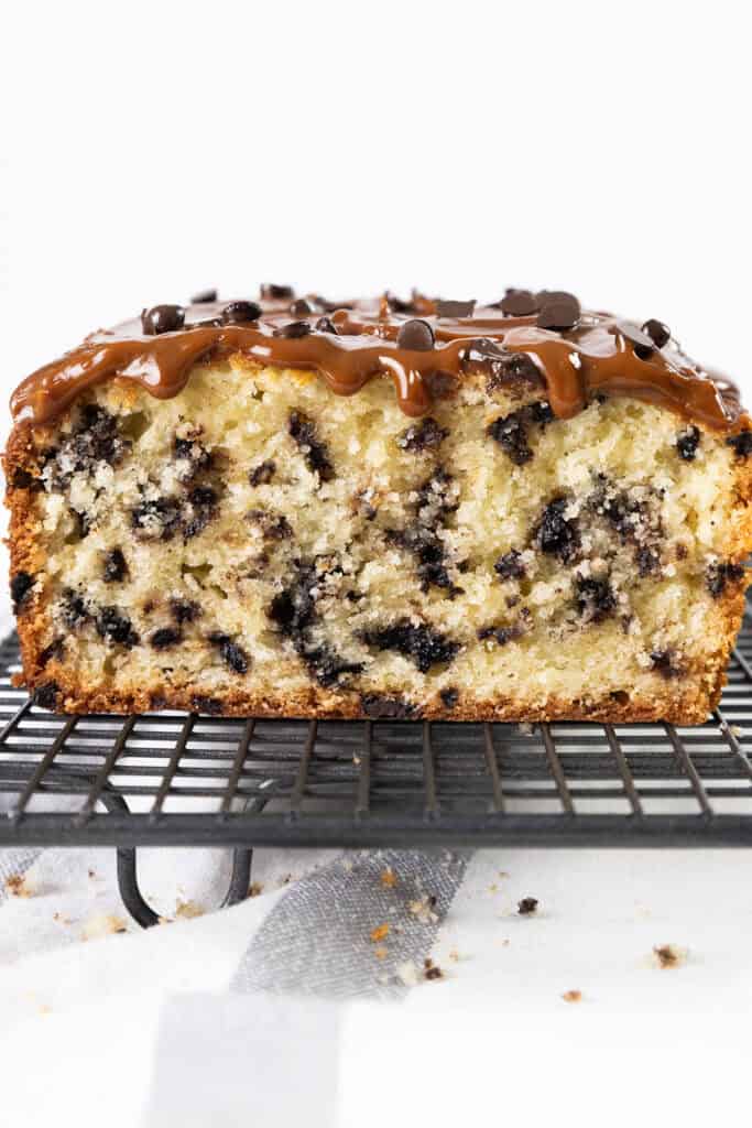 A close-up of a Caramel Chocolate Chip Cake, dripping with rich caramel sauce and sprinkled with chocolate chips. The moist interior is revealed in each slice, dotted generously with chips. It rests invitingly on a cooling rack against a pristine white background.