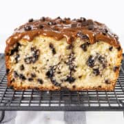 A close-up of a decadent chocolate chip caramel cake studded with chocolate chips. The cake is topped with a glossy chocolate drizzle and extra chips, set on a black wire cooling rack.