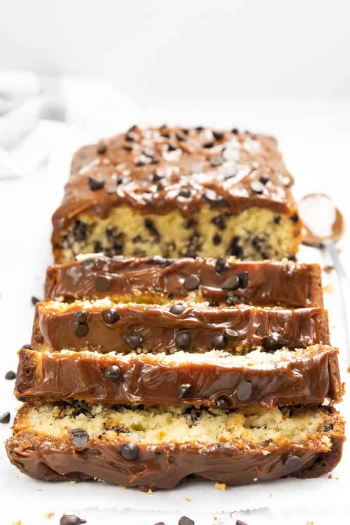 A loaf of chocolate chip caramel cake is sliced, revealing a rich chocolate glaze and sprinkles of chips on top. Inside, pockets of chocolate chips add to the indulgence, with a spoon placed beside it on a pristine white background.