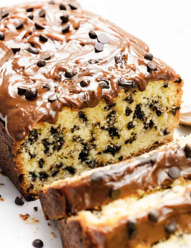 A loaf of Caramel Chocolate Chip Cake topped with melted chocolate and chips. Slices reveal a moist interior, rich with caramel swirls and chocolate chips throughout. It rests elegantly on a white plate.