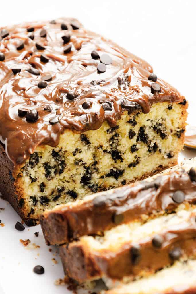 A loaf of Caramel Chocolate Chip Cake topped with melted chocolate and chips. Slices reveal a moist interior, rich with caramel swirls and chocolate chips throughout. It rests elegantly on a white plate.