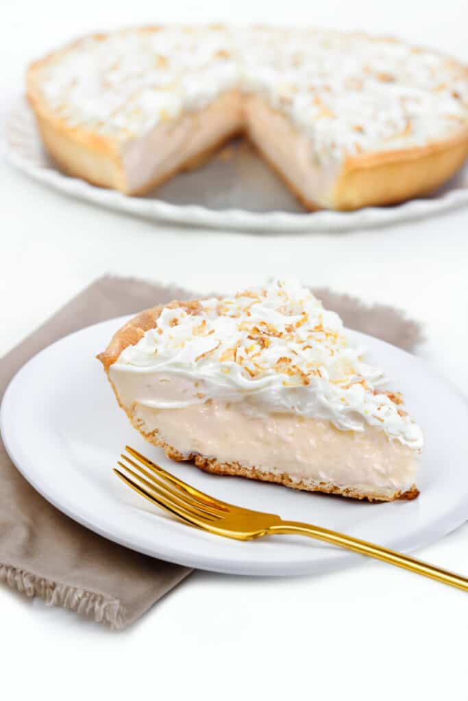 a slice of coconut cream pie on a plate with the pie in the background.