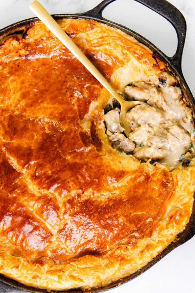 A golden-brown Cheesy Chicken & Mushroom Pie with a flaky crust sits in a cast iron skillet. A wooden spoon has scooped out a portion, revealing a creamy, cheesy filling with chunks of chicken, mushrooms, and vegetables.