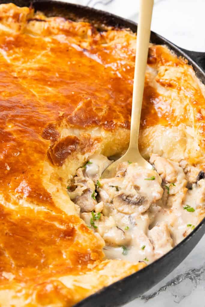 A golden-brown Cheesy Chicken & Mushroom Pie with a flaky crust is shown in a black skillet. A serving spoon reveals the creamy, cheesy filling beneath, dotted with herbs. The pie sits on a marble surface.