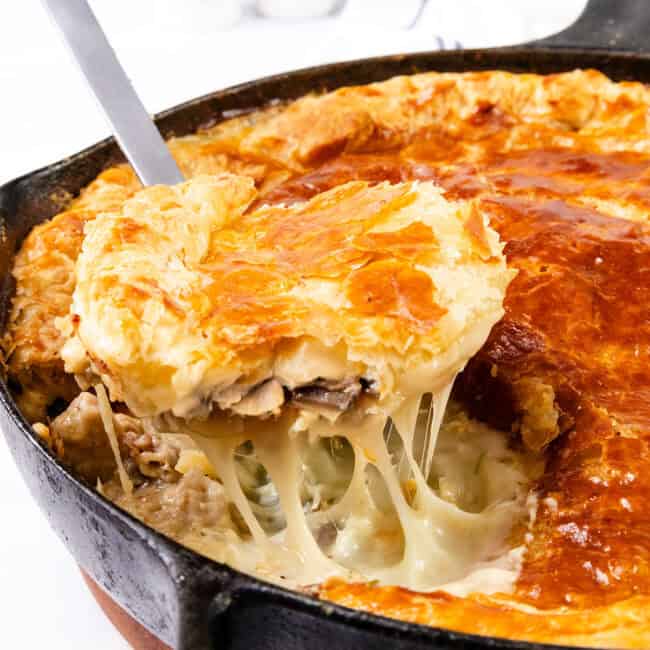 A close-up of a savory Cheesy Chicken & Mushroom Pie in a cast-iron skillet. A piece is being lifted with a serving spatula, revealing a golden, flaky crust and a creamy filling with melted cheese, chicken, mushrooms, and vegetables. Salt and pepper shakers are in the blurred background.