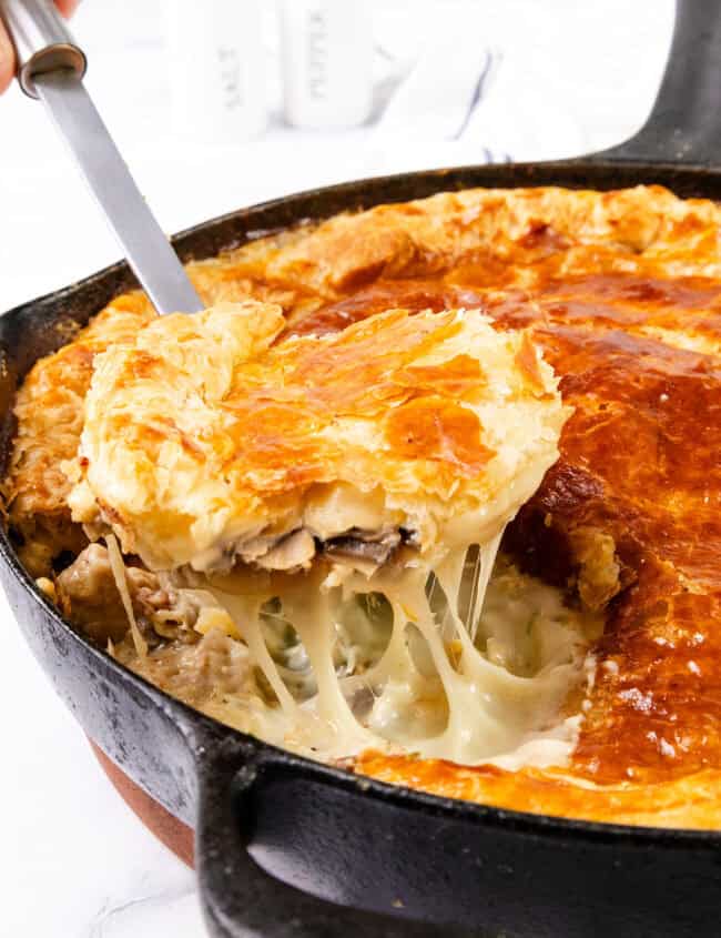 A close-up of a savory Cheesy Chicken & Mushroom Pie in a cast-iron skillet. A piece is being lifted with a serving spatula, revealing a golden, flaky crust and a creamy filling with melted cheese, chicken, mushrooms, and vegetables. Salt and pepper shakers are in the blurred background.