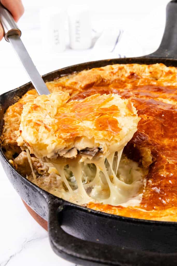 A close-up of a savory Cheesy Chicken & Mushroom Pie in a cast-iron skillet. A piece is being lifted with a serving spatula, revealing a golden, flaky crust and a creamy filling with melted cheese, chicken, mushrooms, and vegetables. Salt and pepper shakers are in the blurred background.