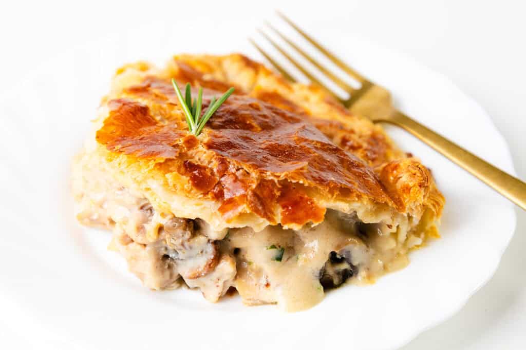 A slice of cheesy chicken and mushroom pie with a golden, flaky crust topped with a rosemary sprig sits on a white plate. A gold fork is placed beside the pie, ready to be used.