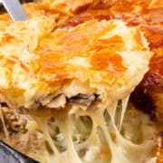 A close-up of a slice being lifted from a Cheesy Chicken & Mushroom Pie. The golden-brown, flaky pastry crust reveals a creamy filling with visible mushrooms and melted cheese strings.